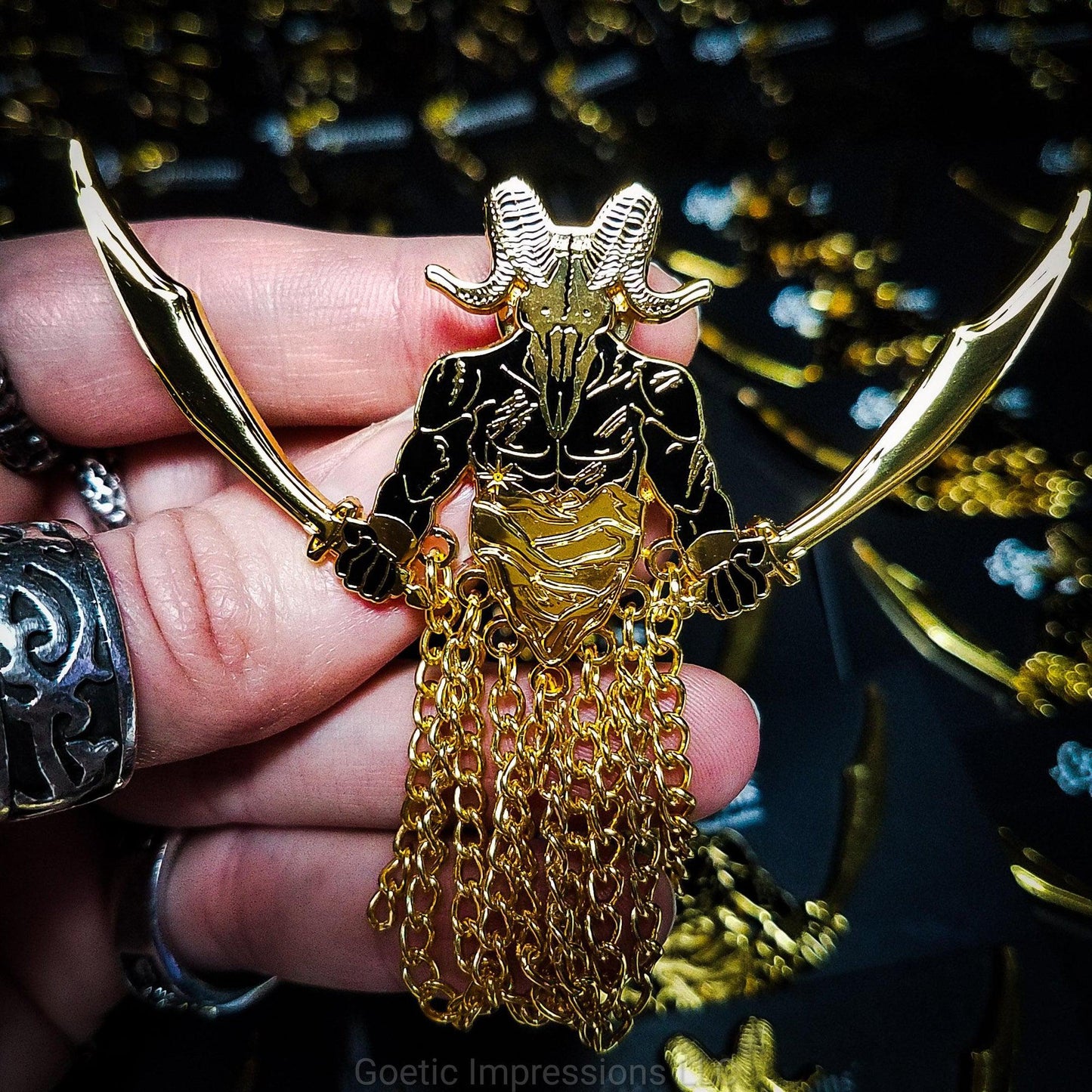 A hand is holding a black and gold hard enamel pin of the demon Azazel. Azazel is shown with the head of a ram skull and human torso brandishing a scimitar in each hand. Azazel is rising up over dunes of a desert. The pin has chains dangling from it.