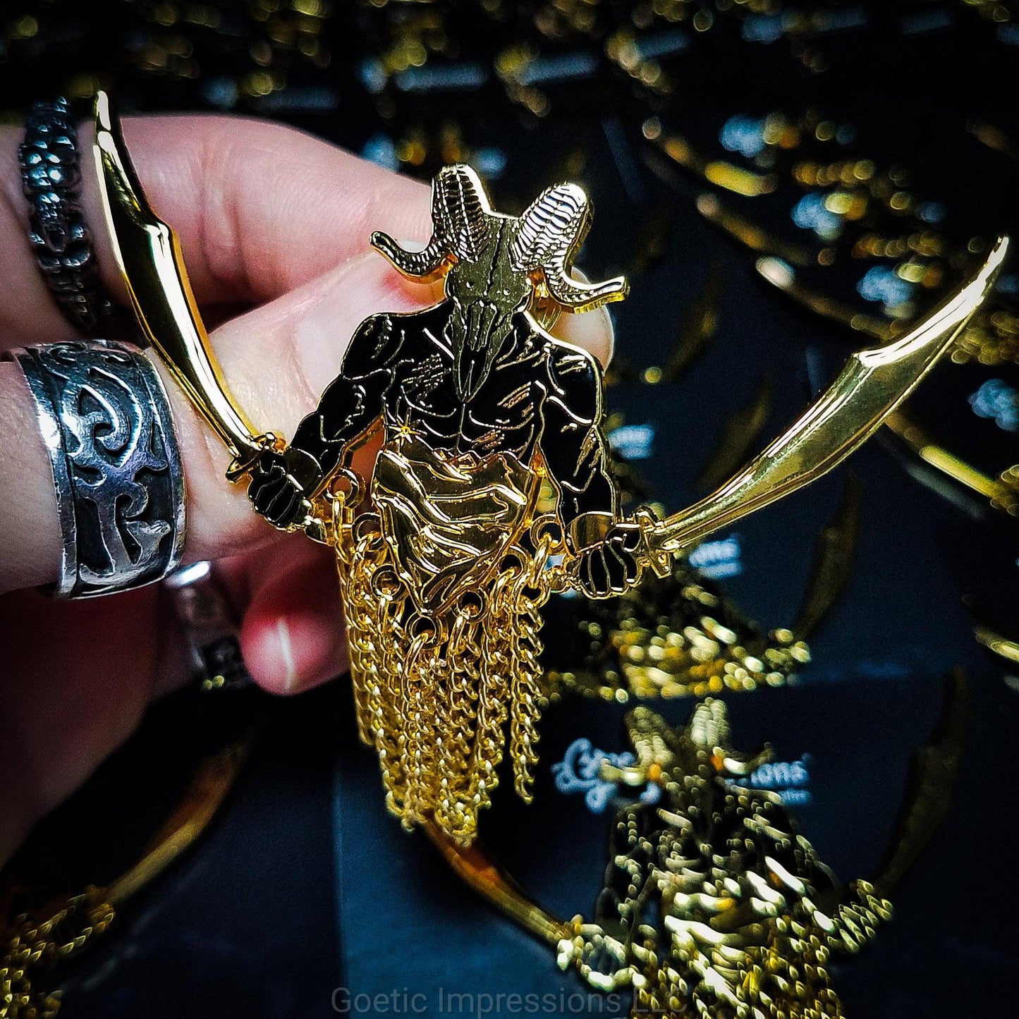A ringed hand holding a black and gold hard enamel pin of the demon Azazel. Azazel is shown with the head of a ram skull and human torso brandishing a scimitar in each hand. Azazel is rising up over dunes of a desert. The pin has chains dangling from it.