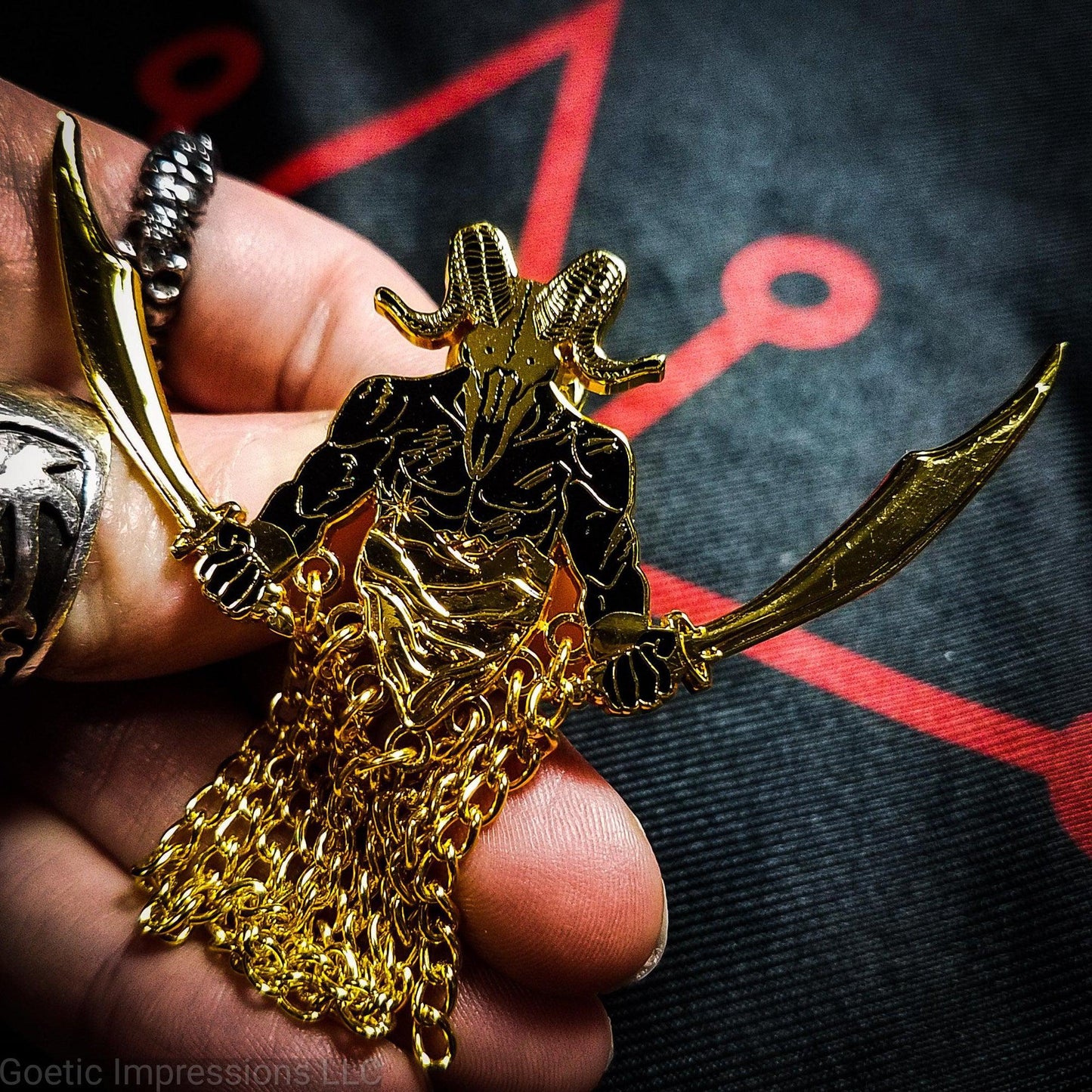 A hand holding a black and gold hard enamel pin of the demon Azazel. Azazel is shown with the head of a ram skull and human torso brandishing a scimitar in each hand. Azazel is rising up over dunes of a desert. The pin has chains dangling from it.