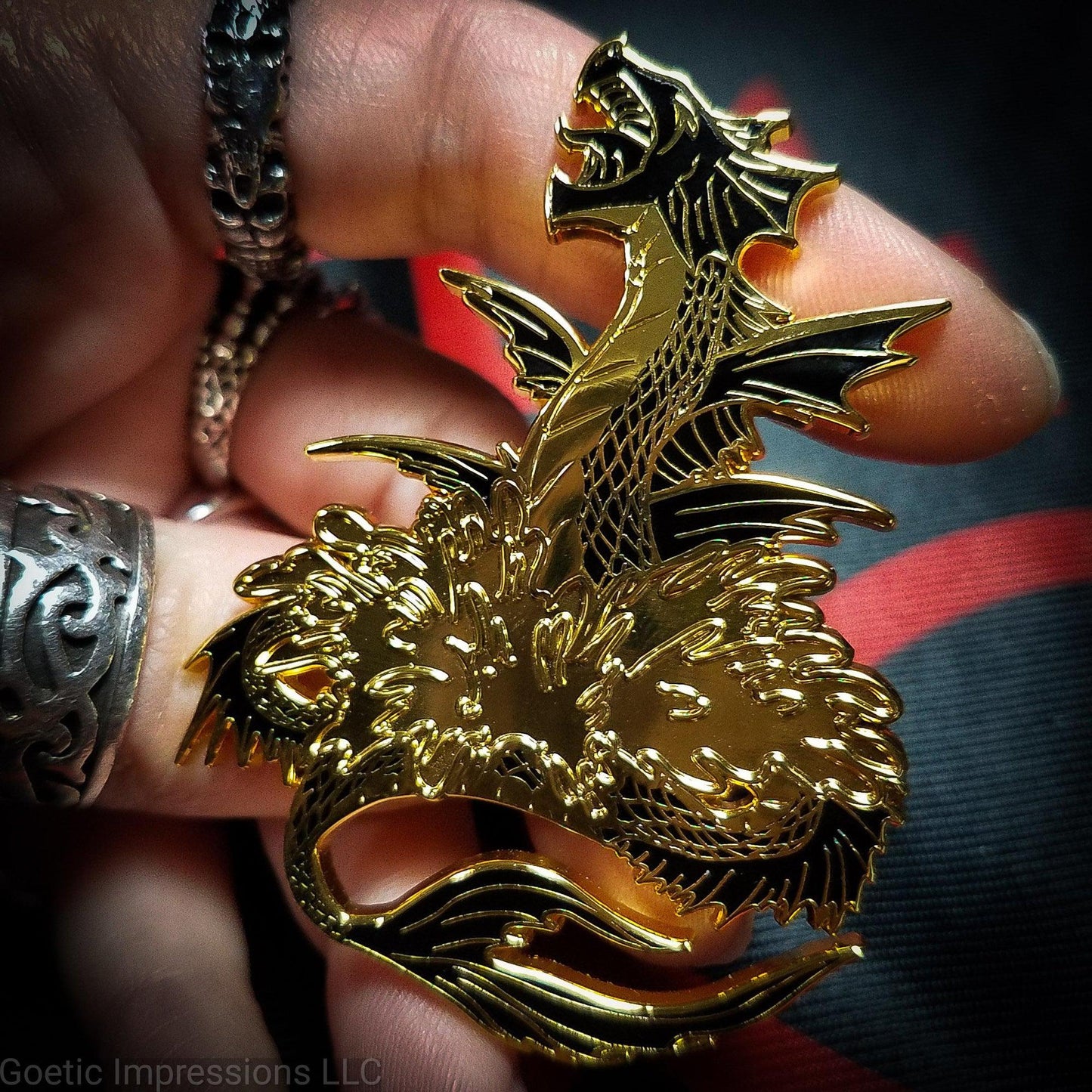 A ringed hand is holding a black and gold hard enamel pin of the sea serpent Leviathan. Leviathan is shown bursting from waters and jaws opened wide.