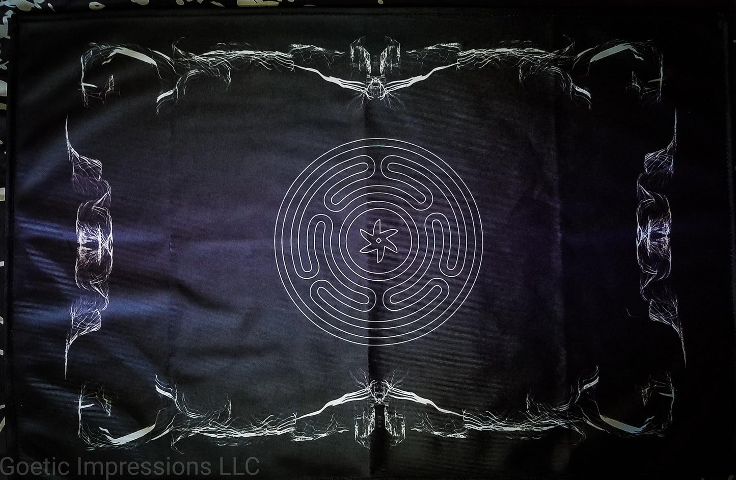 An altar cloth featuring the stropholos or wheel of hecate in the center. The altar cloth background is black and purple with white print and a design around the border. 