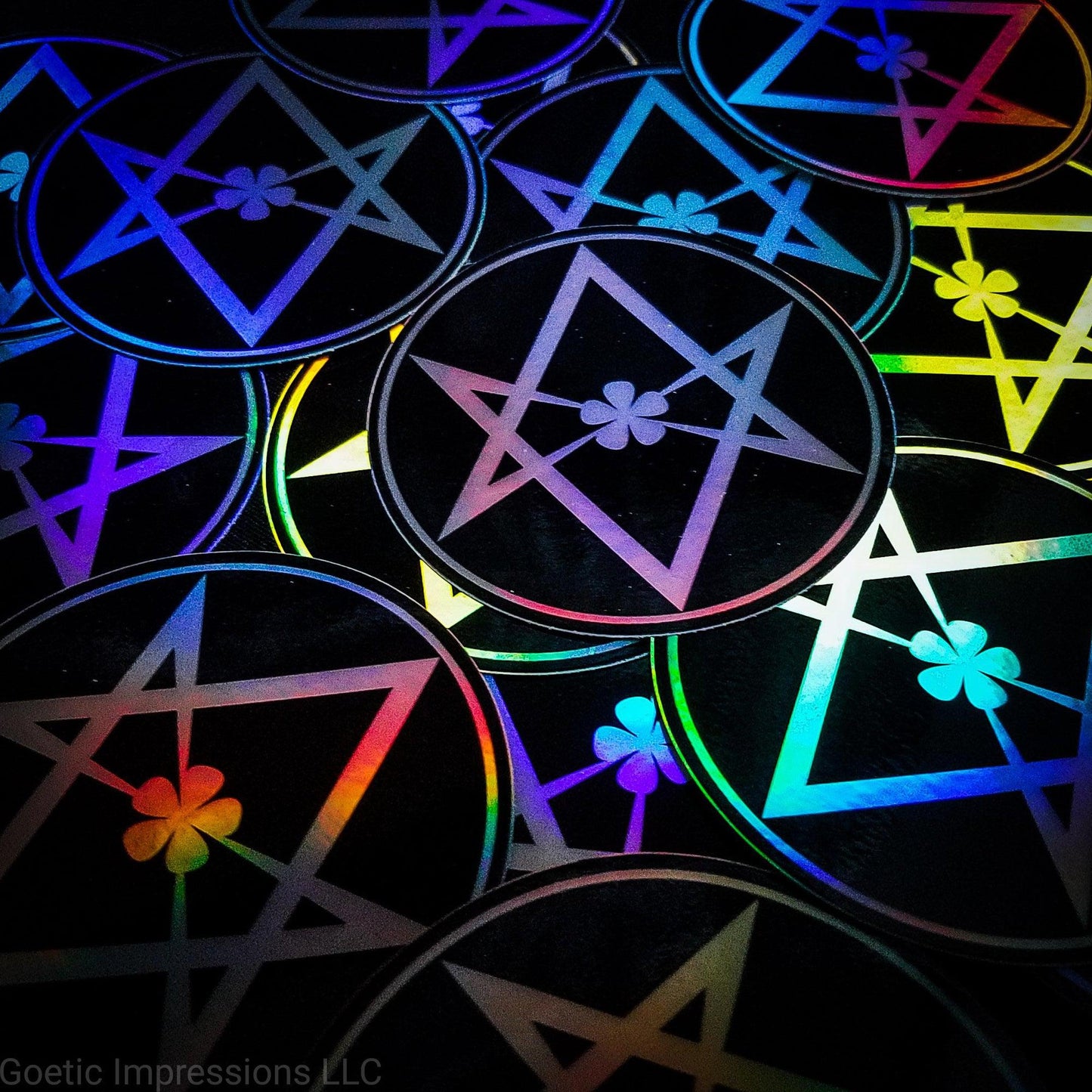 A pile of three inch circular holographic stickers. The stickers have a black background and a white symbol featuring the Unicursal Hexagram with a flower petal in the center. The holographic paper shines in a wide arrange of rainbow colors when the light catches it at different angles.