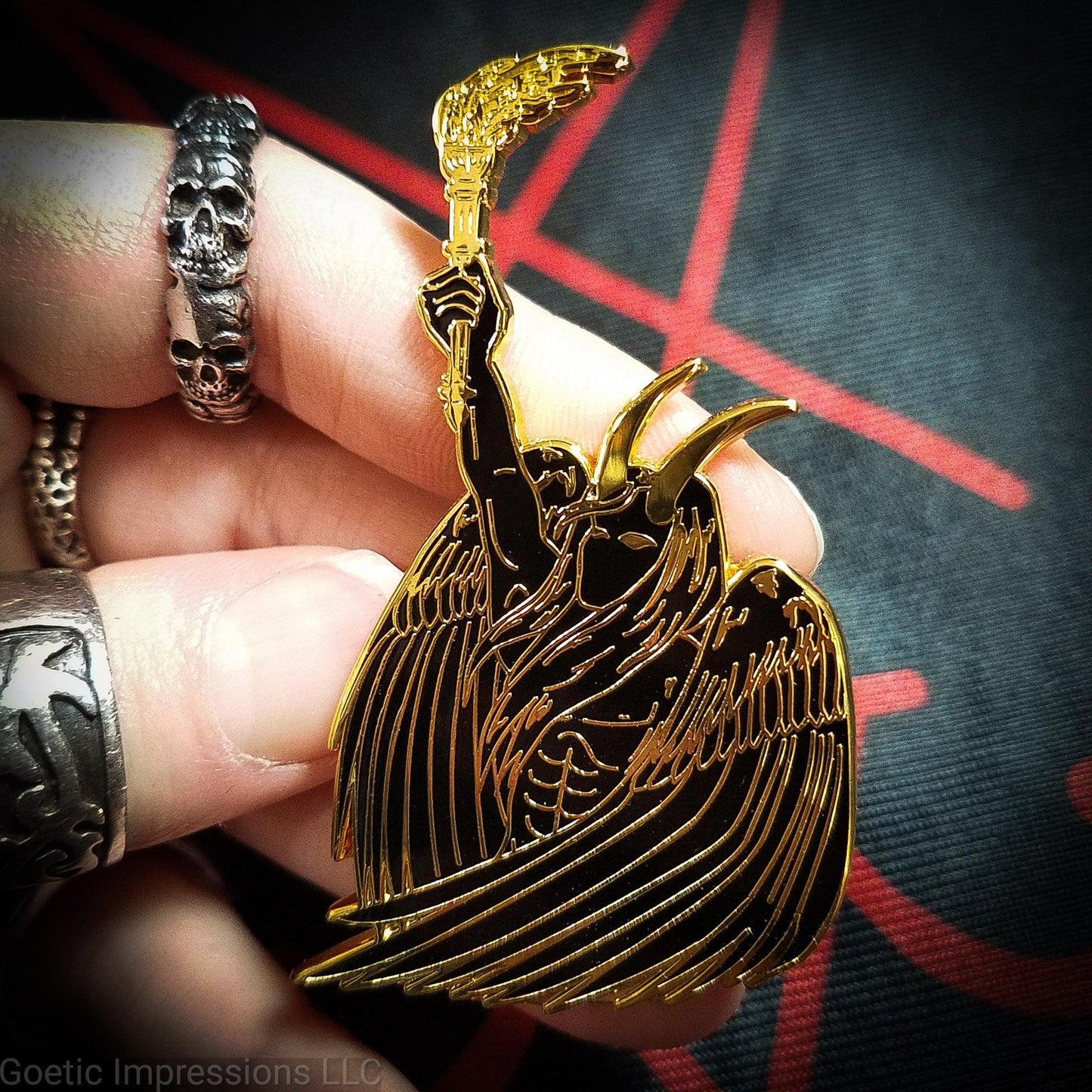 A ringed hand is holding a black and gold hard enamel pin of Lucifer. Lucifer is holding a flaming torch held high and has his wings surrounding him. He has horns and is bare chested.