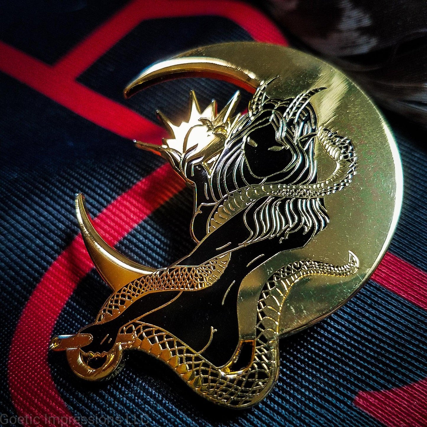 A black and gold hard enamel pin of Lilith. Lilith is holding a shining apple in one hand and a rod and circle in the other. Her one arm is coiled with a serpent. A cresent moon is behind her.