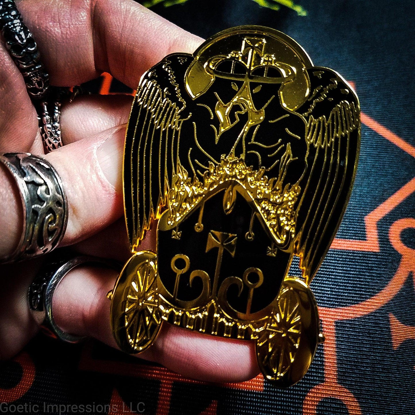 A hand is holding a black and gold hard enamel pin featuring the demon Belial. Belial is shown as two angels facing each other with arms interlocked. They are standing in a flaming chariot.