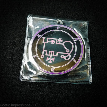 Amulet of Phenex in a PVC pouch. The sigil for Phenex is pink. Phenex's name is surrounding the sigil with concentric circles in purple on a black background. The seal is silver plated.
