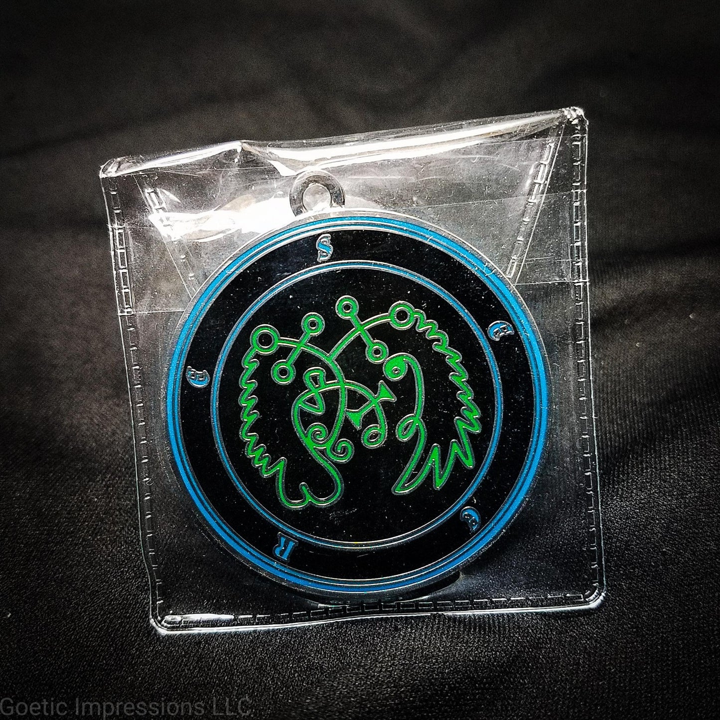 Amulet of Seere in a PVC pouch. The sigil for Seere is green. Seere's name is surrounding the sigil with concentric circles in blue on a black background.  The seal is silver plated.