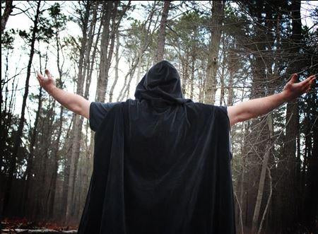 A cloaked magician facing away looking at trees with arms outspread in adoration.