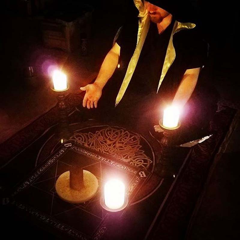 Ceremonial magician kneeled at an Enochian altar with the Table of Practice and a Wax Sigillum Dei Aemeth in the center. On top of the Sigillum is a quartz pillar. At the four corners of the table are lit candles.