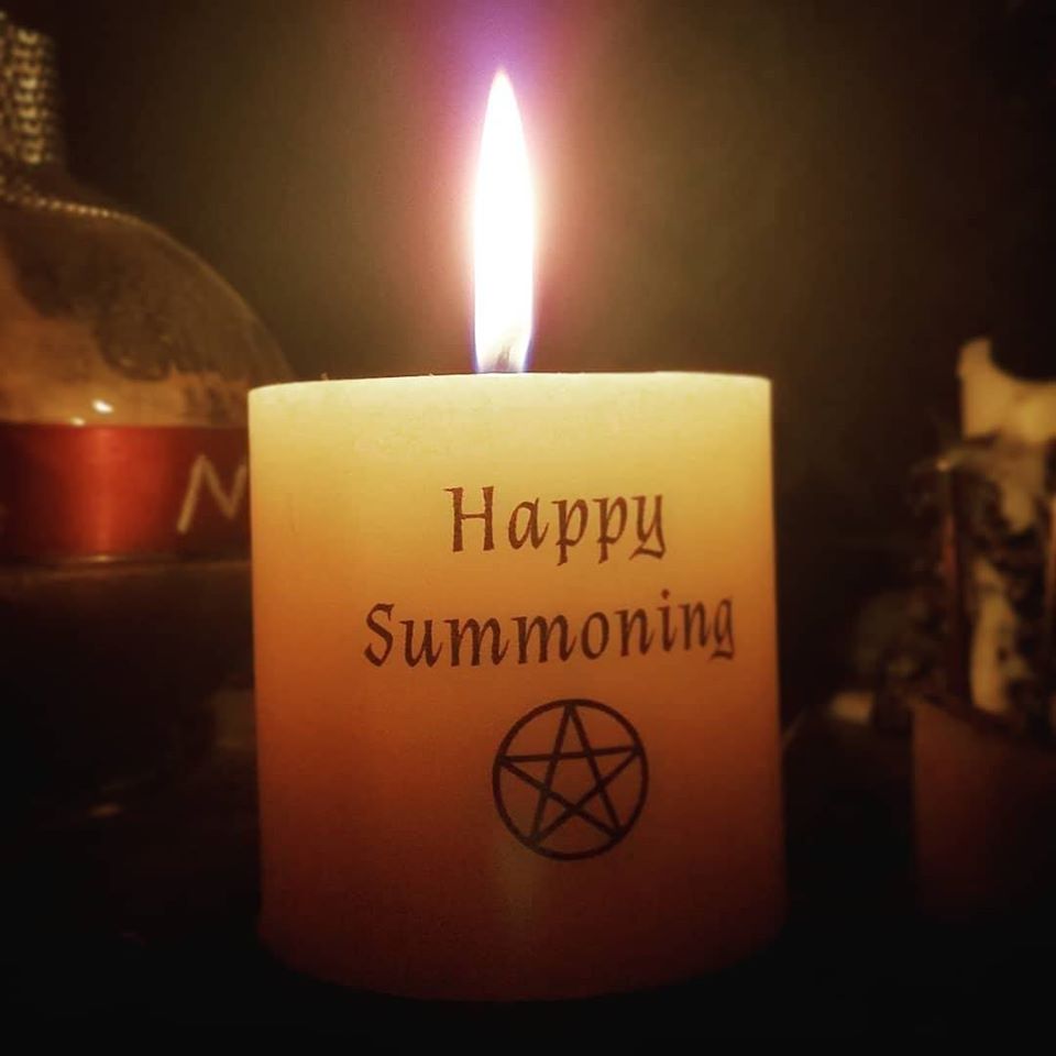 A lit candle with a burning flame on an altar. The candle is imprinted with the words "Happy Summoning" with a pentacle beneath the letters.