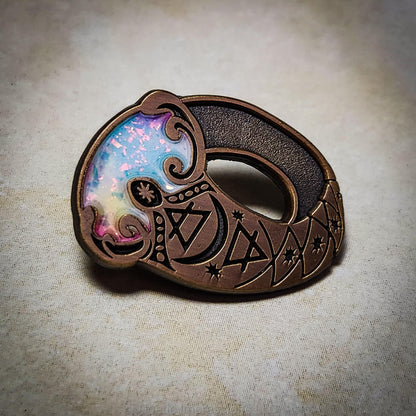 Enamel pin of a fairy ring of invisiblity in antique gold enamel and a color sparkly opal center. The ring is engraved with Moon, Air and Earth alchemical symbols.