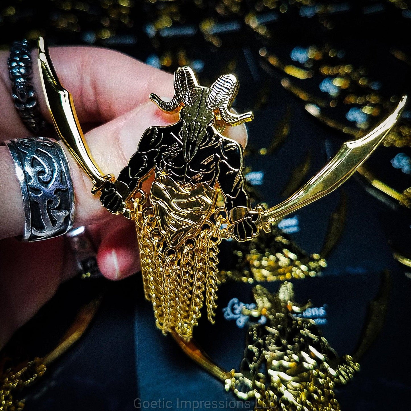 A ringed hand holding a black and gold hard enamel pin of the demon Azazel. Azazel is shown with the head of a ram skull and human torso brandishing a scimitar in each hand. Azazel is rising up over dunes of a desert.  The pin has chains dangling from it. 