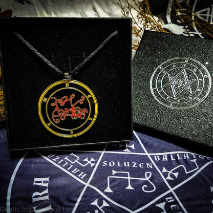 Necklace of Beleth in a Goetic Impressions gift box stamped in silver foil. The sigil for Beleth is red. Beleth's name is surrounding the sigil with concentric circles in yellow on a black background. The seal is gold plated. The box is on a purple altar cloth with the Tetragrammaton in white.
