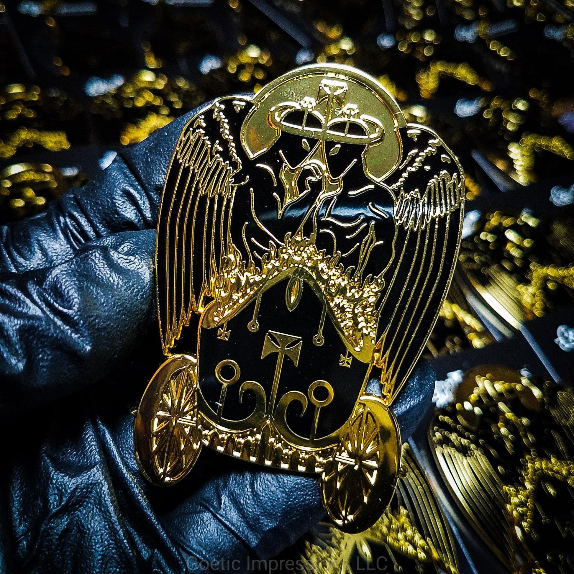 A gloved hand is holding a hard enamel pin that is black and gold featuring the demon king, Belial. Belial is shown as two twin angels facing each other with an arm interlocked with the other.  The are standing in a chariot of fire.