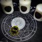 A black and white altar cloth with a seal of the planetary Archangels in the center. The seal contains various astrological sigils. There are candles and other ritual tools on the cloth.