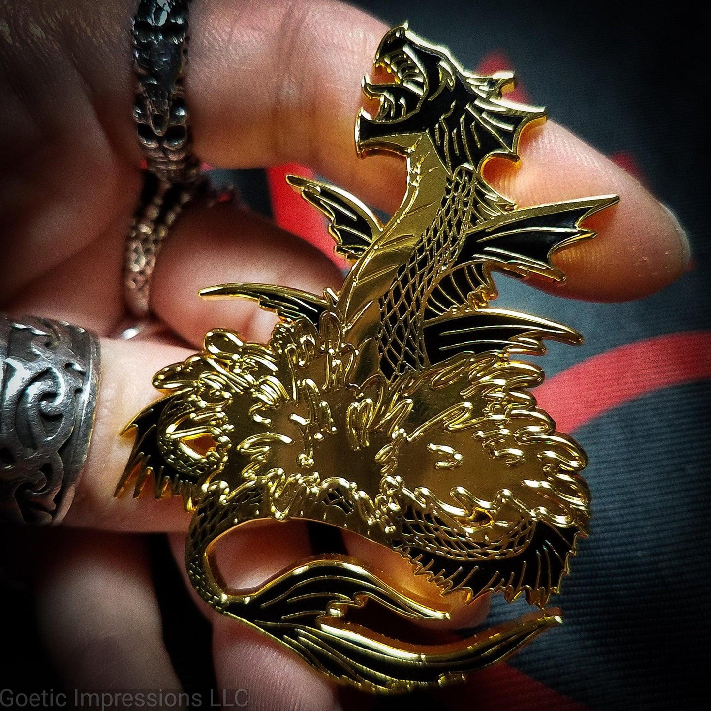 A ringed hand is holding a black and gold hard enamel pin of the sea serpent Leviathan. Leviathan is shown bursting from waters and jaws opened wide. 