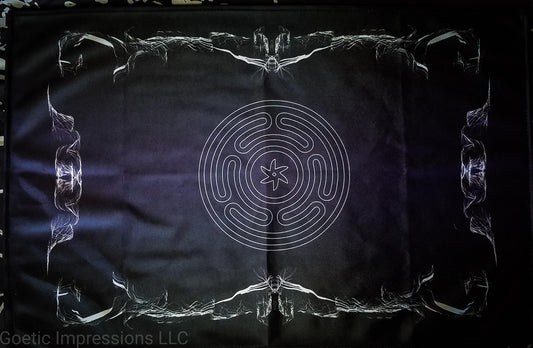 An altar cloth featuring the stropholos or wheel of hecate in the center. The altar cloth background is black and purple with white print and a design around the border. 
