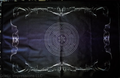 An altar cloth featuring the stropholos or wheel of hecate in the center. The altar cloth background is black and purple with white print and a design around the border. 