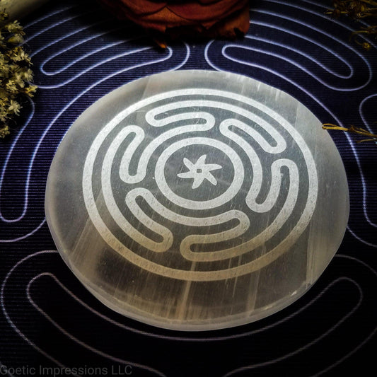 A selenite charging plate with the Wheel of Hecate or Stropholos engraved in it. The plate is placed on a purple altar cloth also with a white hecate sigil.