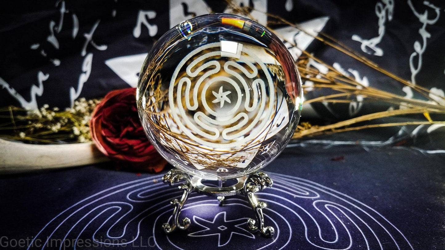 A crystal ball with the engraved sigil of Hecate in the center. The ball sits on a silver stand. This sigil is also called the Stropholos or Wheel of Hecate.. The crystal ball is on an altar cloth with the same sigil.