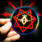 A hand holding a three inch circular holographic sticker. The sticker has a black background and a white and red symbol featuring the Star of Babalon. The holographic paper shines in a wide arrange of multicolors when the light catches it.