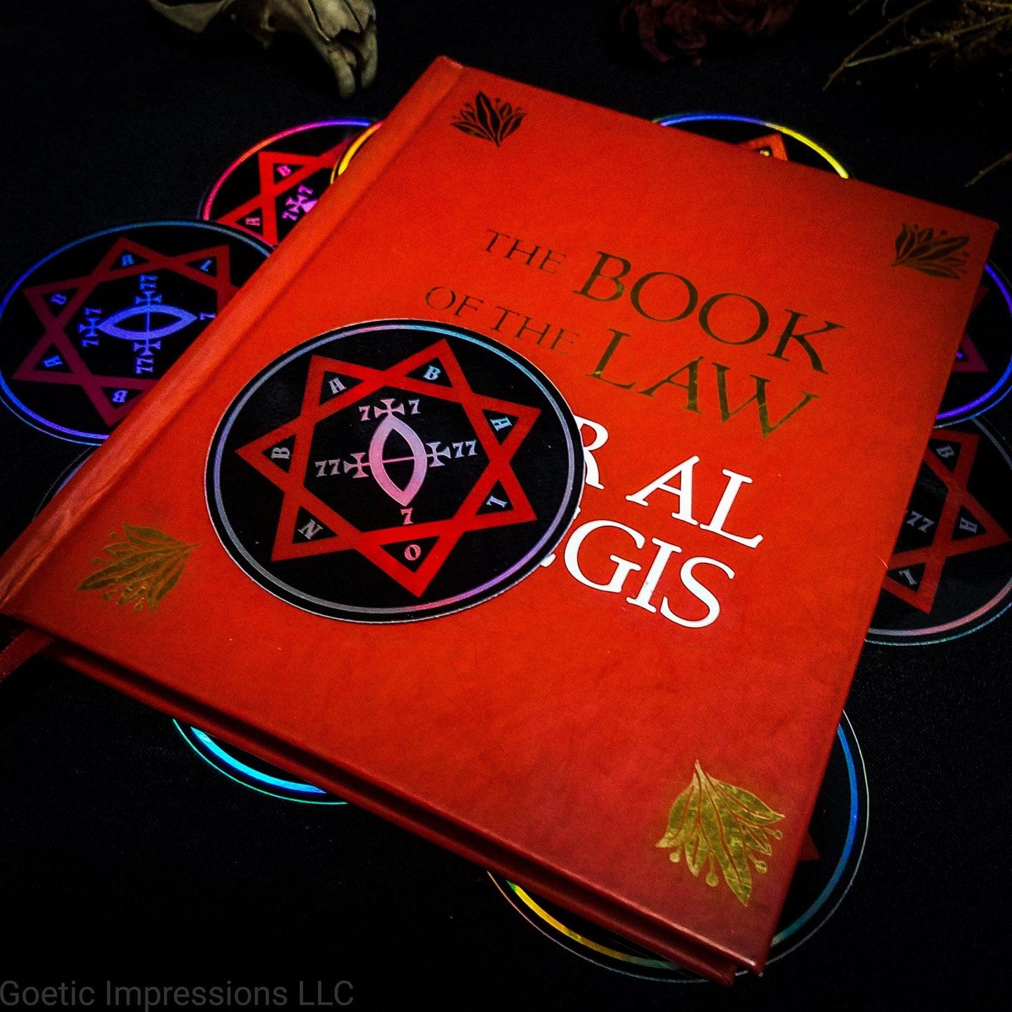 A three inch holographic sticker with a black background and a white and red symbol of a Star of Babalon rests on top of a red book titled Book of the Law, Liber Al Vel Legis. The book is placed on a pile of the stickers. The holographic paper shines in a wide arrange of rainbow colors when the light catches it at different angles.