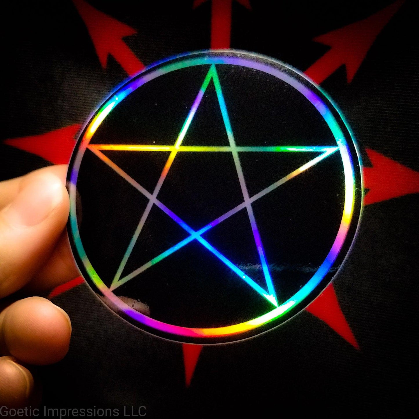 A hand holding a three inch circular holographic sticker. The sticker has a black background and a white symbol featuring a pentacle. The holographic paper shines in a wide arrange of multicolors when the light catches it.