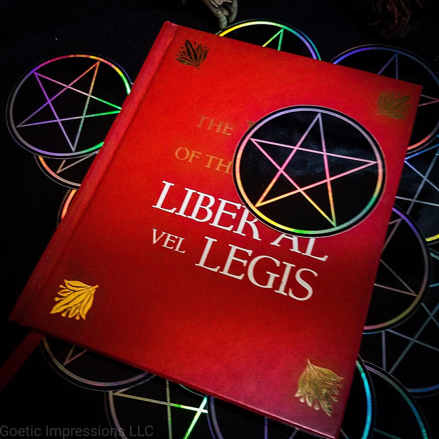 A three inch holographic sticker with a black background and a white symbol of a Pentacle rests on top of a red book titled Book of the Law, Liber Al Vel Legis. The book is placed on a pile of the stickers. The holographic paper shines in a wide arrange of rainbow colors when the light catches it at different angles.