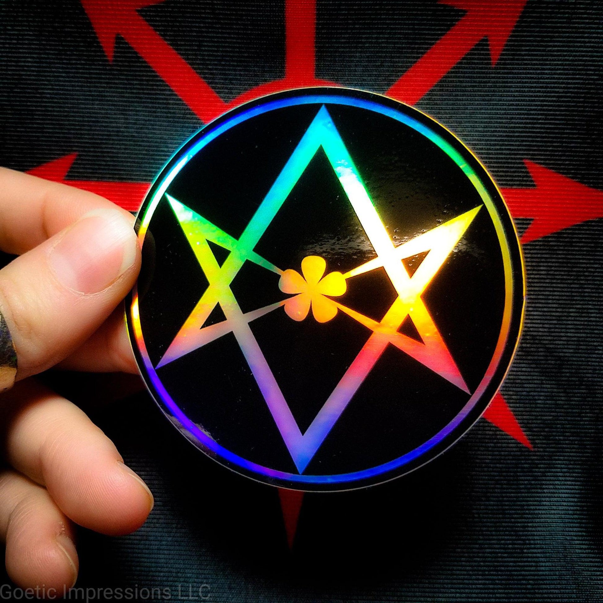 A hand holding a three inch circular holographic sticker. The sticker has a black background and a white symbol featuring the Unicursal Hexagram with a flower petal in the center. The holographic paper shines in a wide arrange of multicolors when the light catches it.