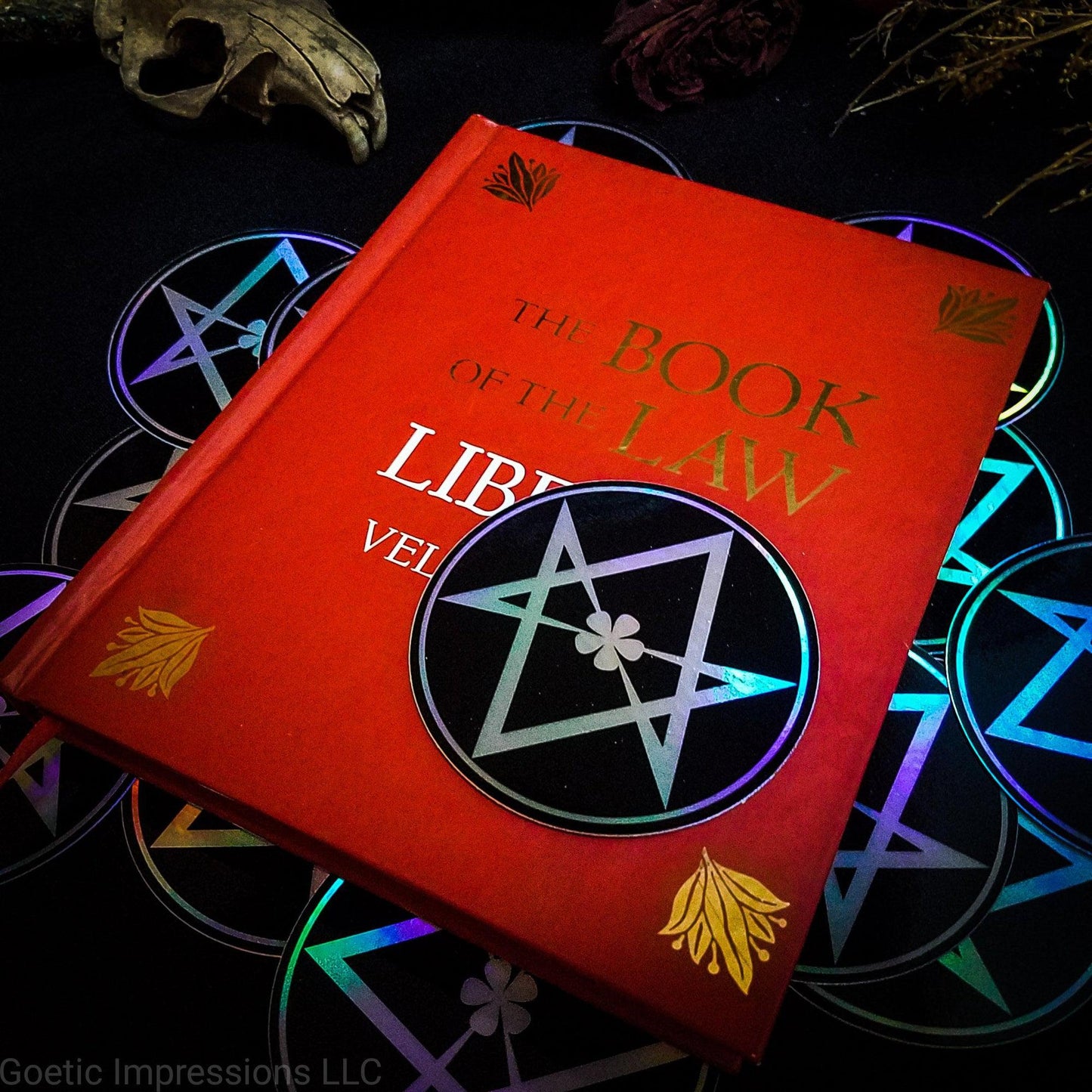 A three inch holographic sticker with a black background and a white symbol of a Unicursal Hexagram rests on top of a red book titled Book of the Law, Liber Al Vel Legis. The book is placed on a pile of the stickers. The holographic paper shines in a wide arrange of rainbow colors when the light catches it at different angles.