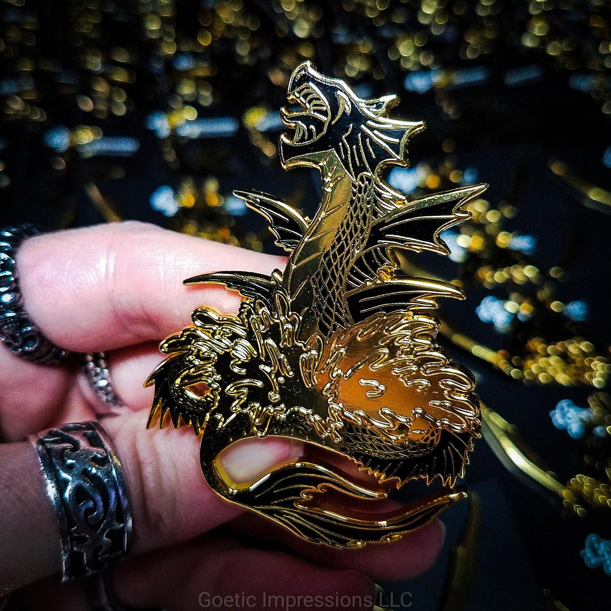 A ringed hand holding a black and gold hard enamel pin of the sea serpent Leviathan. Leviathan is shown bursting from waters and jaws wide open. 