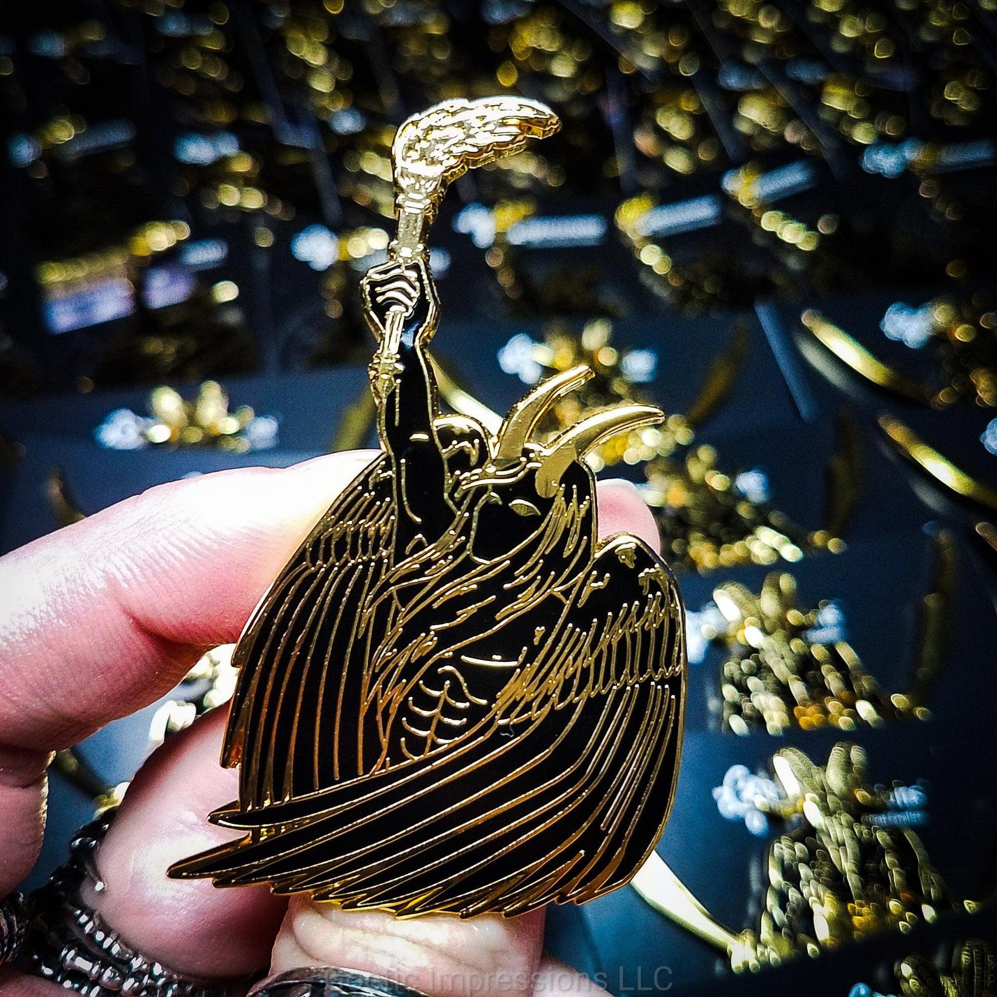 A ringed hand is holding a black and gold hard enamel pin of Lucifer. Lucifer is holding a flaming torch held high and has his wings surrounding him. He has horns and is bare chested.