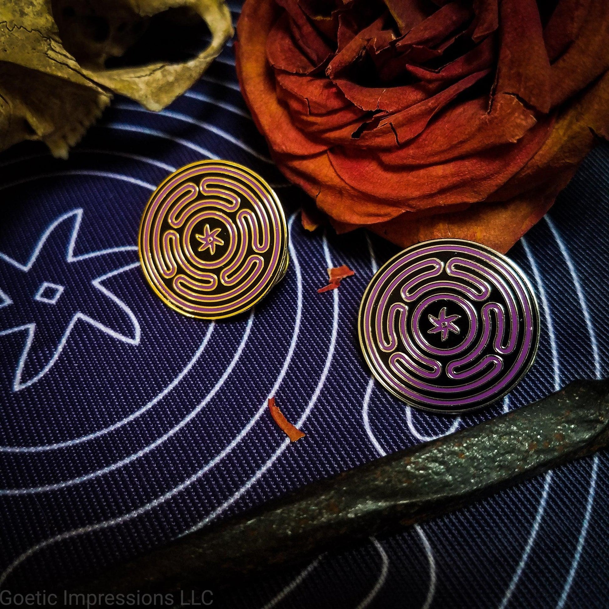Two hard enamel pins featuring the Strophlos or Wheel of Hecate sigil. The sigil itself is colored in purple on a black background. The pins come in silver or gold plating. The pins are on an altar cloth with the same sigil surrounded by ritual tools.