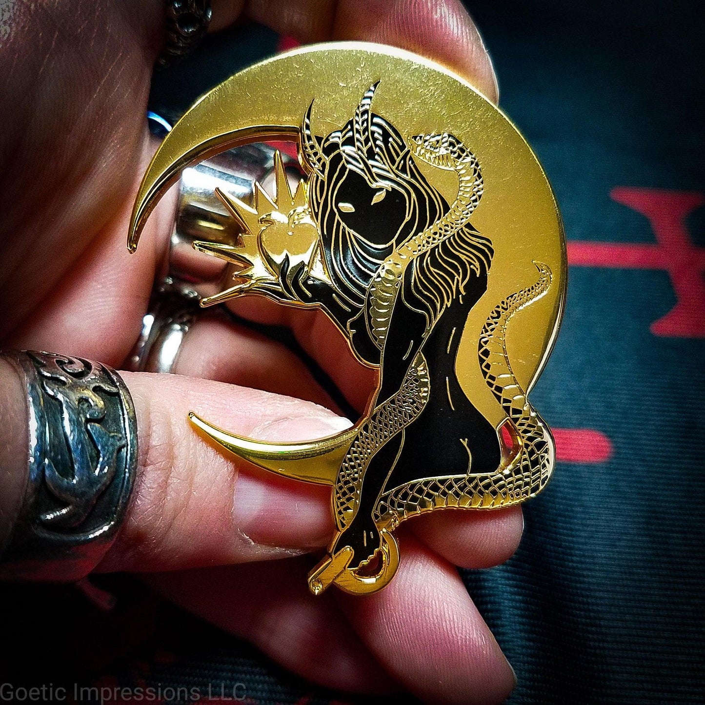 A hand holding a black and gold hard enamel pin of Lilith. Lilith is holding a shining apple in one hand and a rod and circle in the other. Her one arm is coiled with a serpent. A cresent moon is behind her.