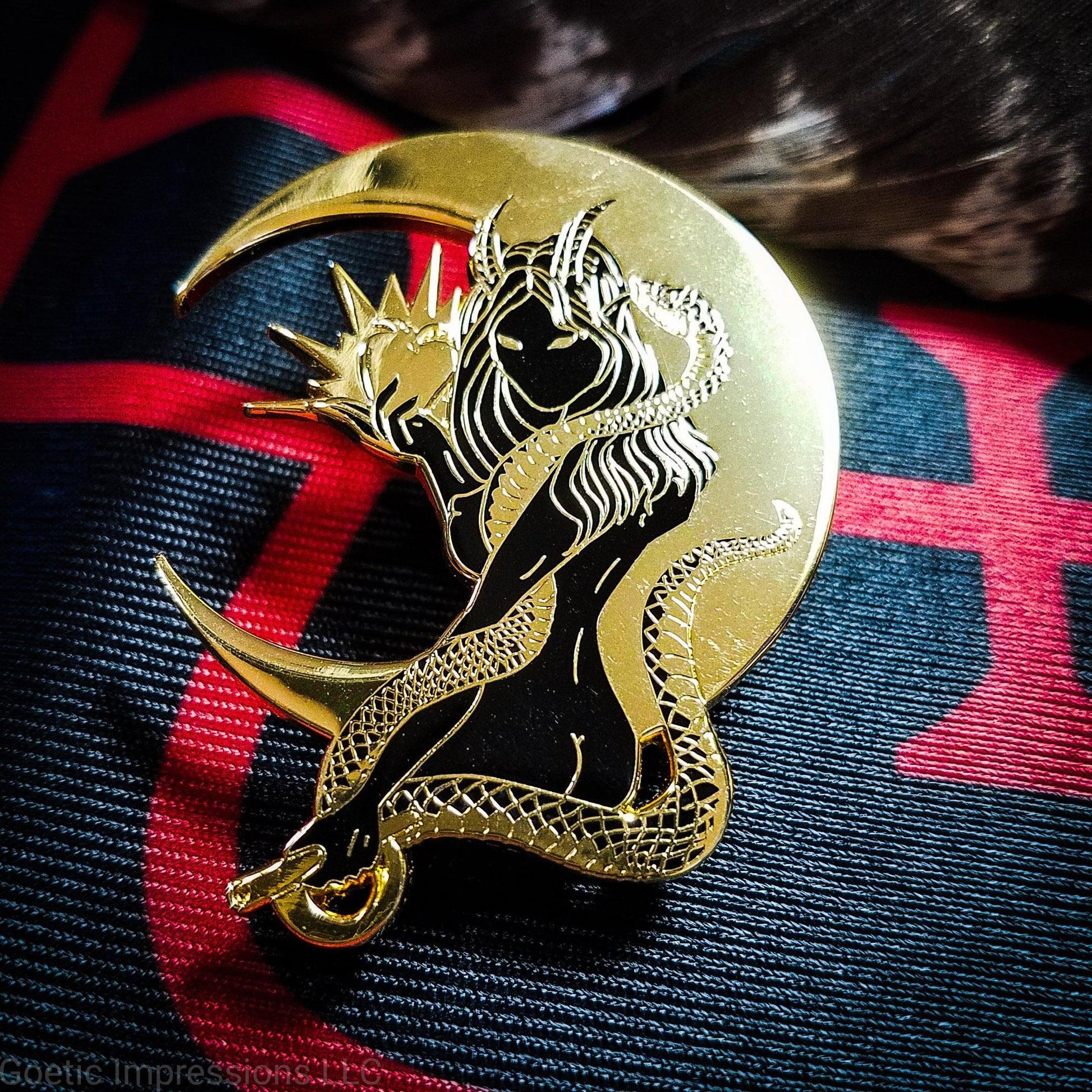 A black and gold hard enamel pin of Lilith. Lilith is holding a shining apple in one hand and a rod and circle in the other. Her one arm is coiled with a serpent. A cresent moon is behind her.