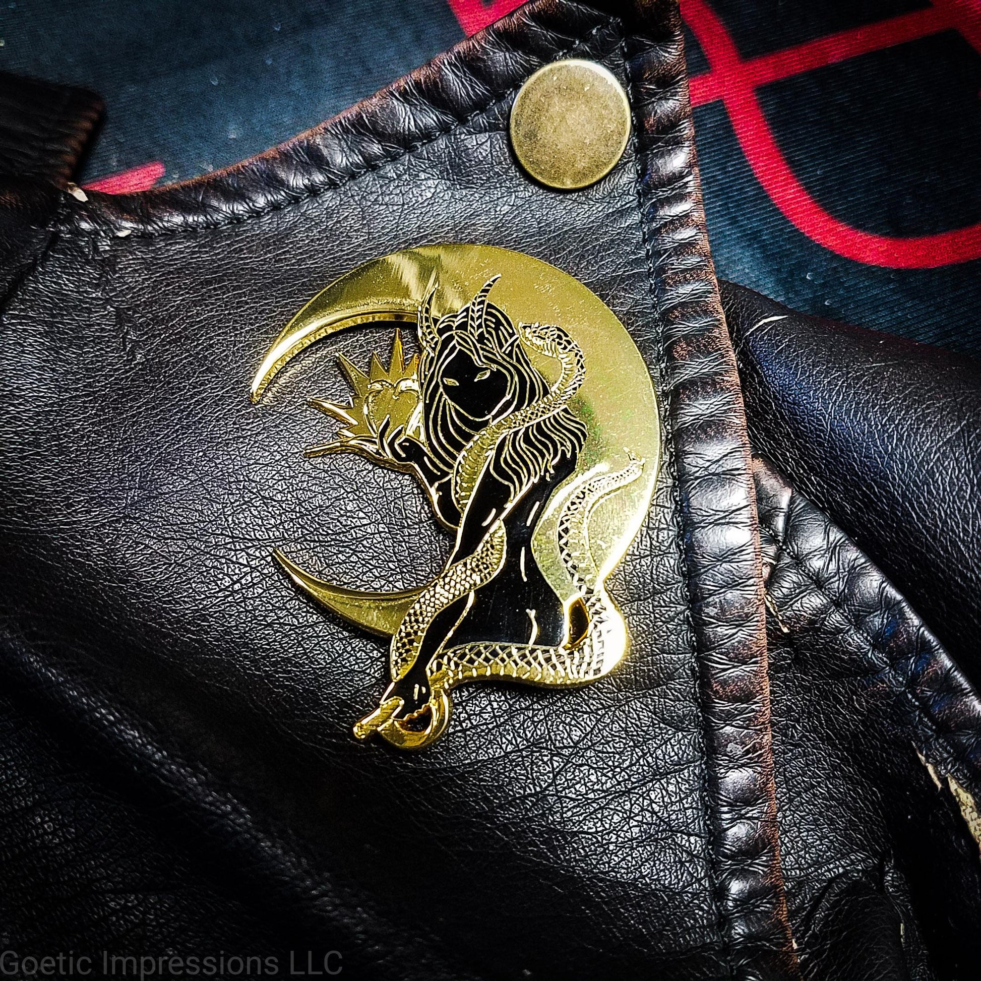 A black and gold hard enamel pin of Lilith. Lilith is holding a shining apple in one hand and a rod and circle in the other. Her one arm is coiled with a serpent. A cresent moon is behind her. The pin is on a brown leather jacket.