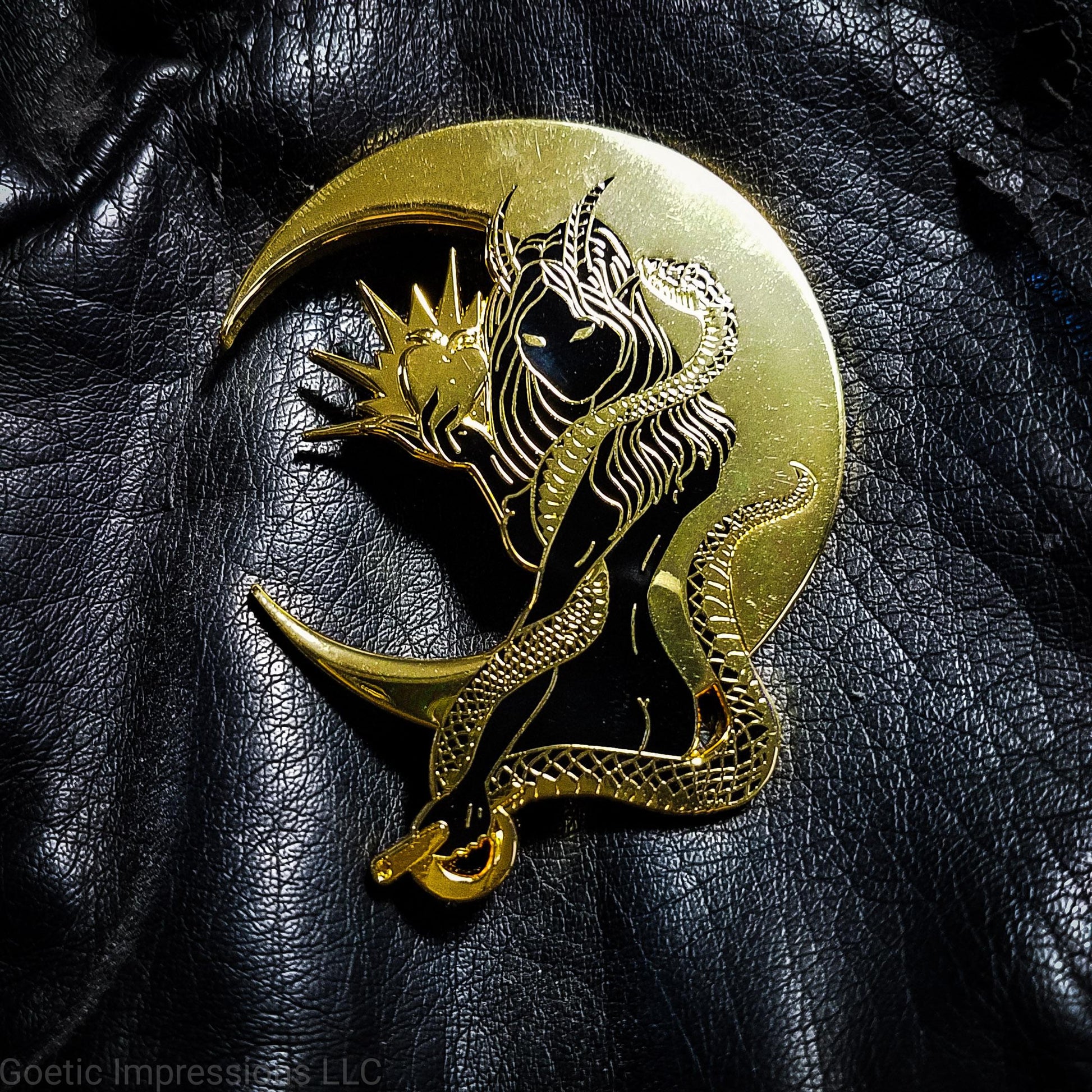 A black and gold hard enamel pin of Lilith. Lilith is holding a shining apple in one hand and a rod and circle in the other. Her one arm is coiled with a serpent. A cresent moon is behind her. The pin is on a black leather jacket