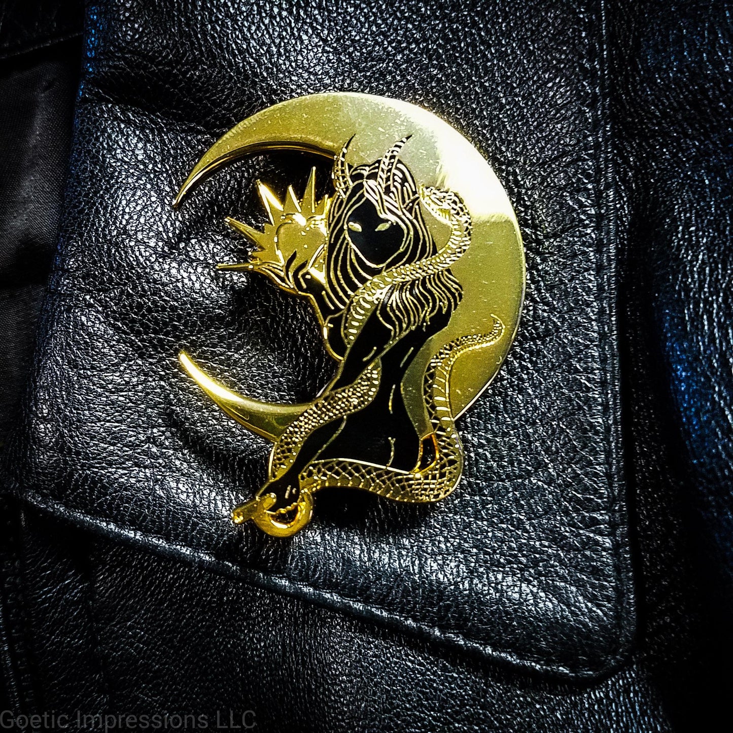 A black and gold pin of the demoness Lilith on a black leather jacket. The hard enamel pin shows Lilith holding a glowing apple riased in one hand and a serpent encricling her other arm. A cresent moon is backdropped behind her. She is also holding a circle and rod in her other hand.