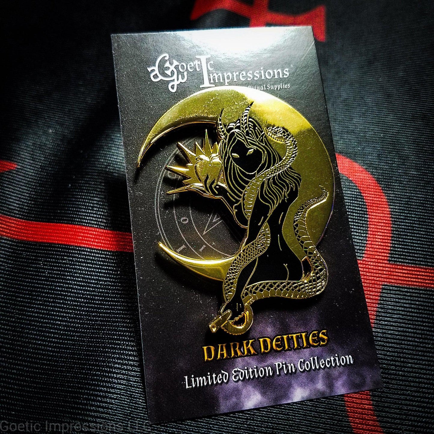 A black and gold hard enamel pin of Lilith. Lilith is holding a shining apple in one hand and a rod and circle in the other. Her one arm is coiled with a serpent. A cresent moon is behind her. The pin is positioned on a Goetic Impressions card back.