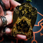 A hand is holding a black and gold hard enamel pin featuring the demon Belial. Belial is shown as two angels facing each other with arms interlocked.  They are standing in a flaming chariot. 