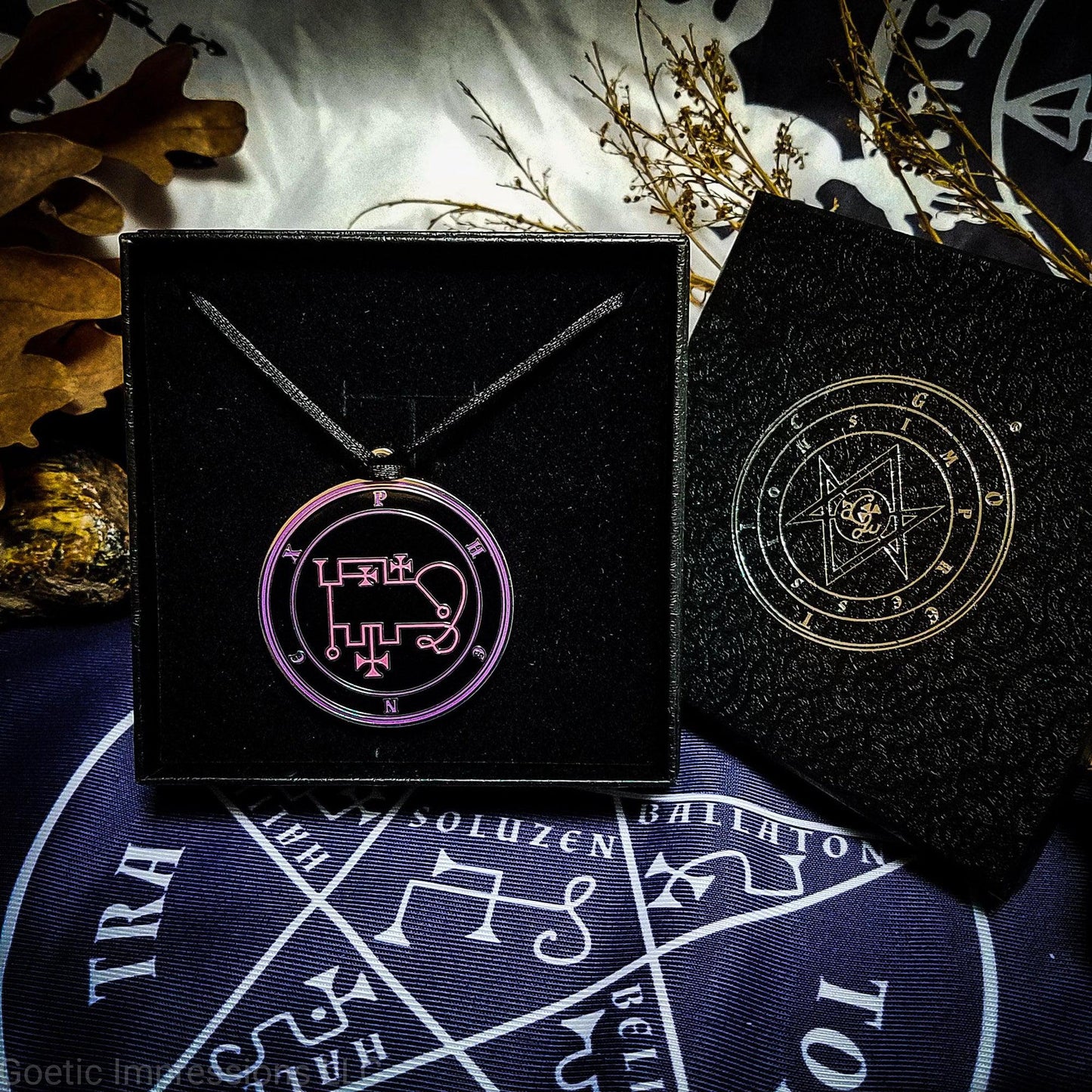 Necklace of Phenex in a Goetic Impressions gift box stamped in silver foil. The sigil for Phenex is pink. Phenex's name is surrounding the sigil with concentric circles in purple on a black background. The seal is silver plated. The box is on a purple altar cloth with the Tetragrammaton in white.