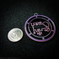 Seal of Phenex next to a quarter. The seal medallion measures two inches in diameter. The sigil is pink with purple letters and circles on a black background. 