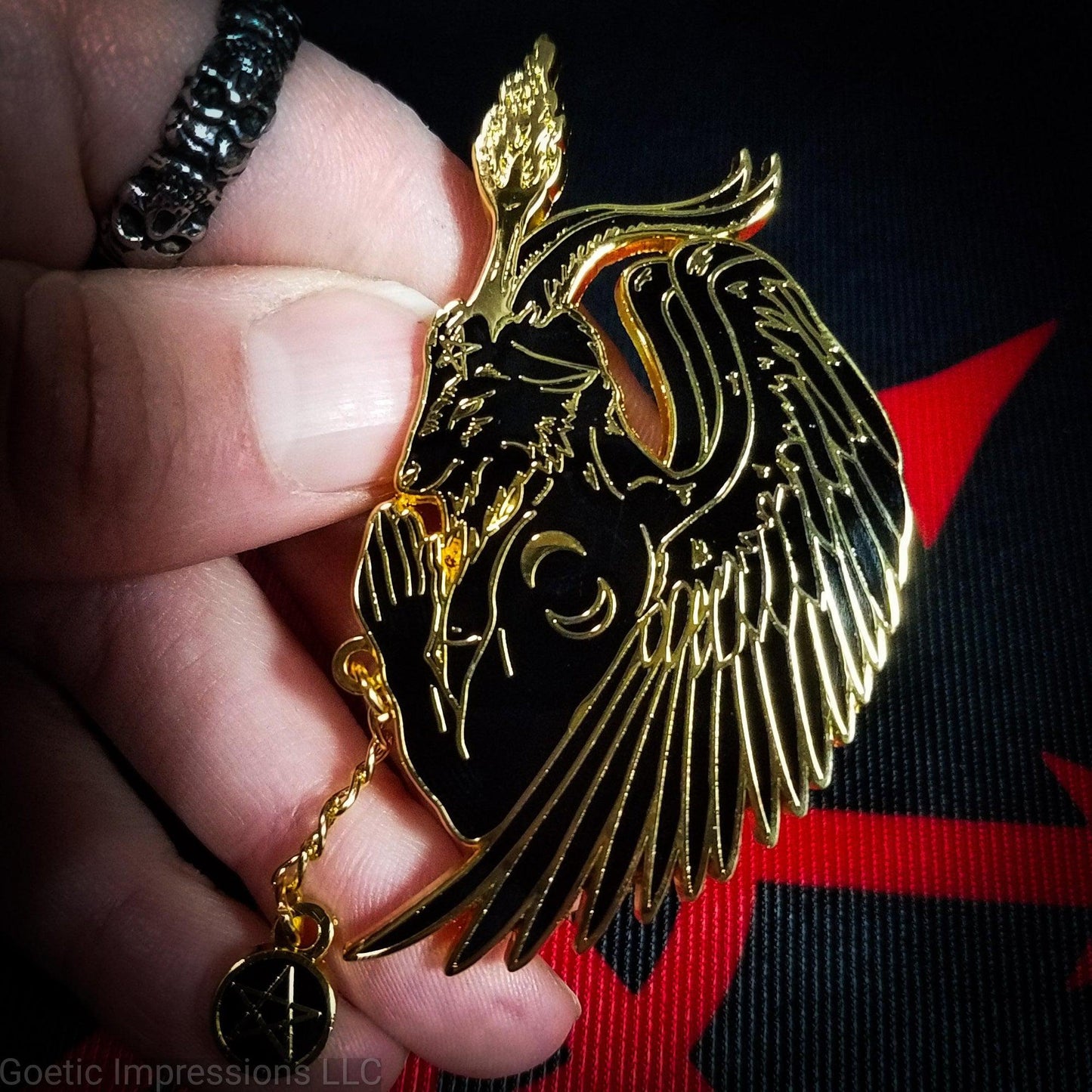 A hand holding a black and gold hard enamel pin featuring Baphomet in a side profile praying pose. There is a pentacle chain rosary in Baphomet's hands.