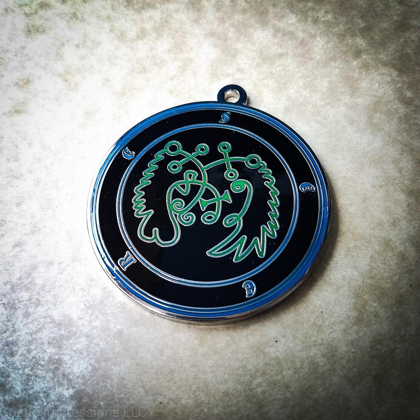 Amulet of Seere. The sigil for Seere is green. Seere's name is surrounding the sigil with concentric circles in blue on a black background.  The seal is silver plated.