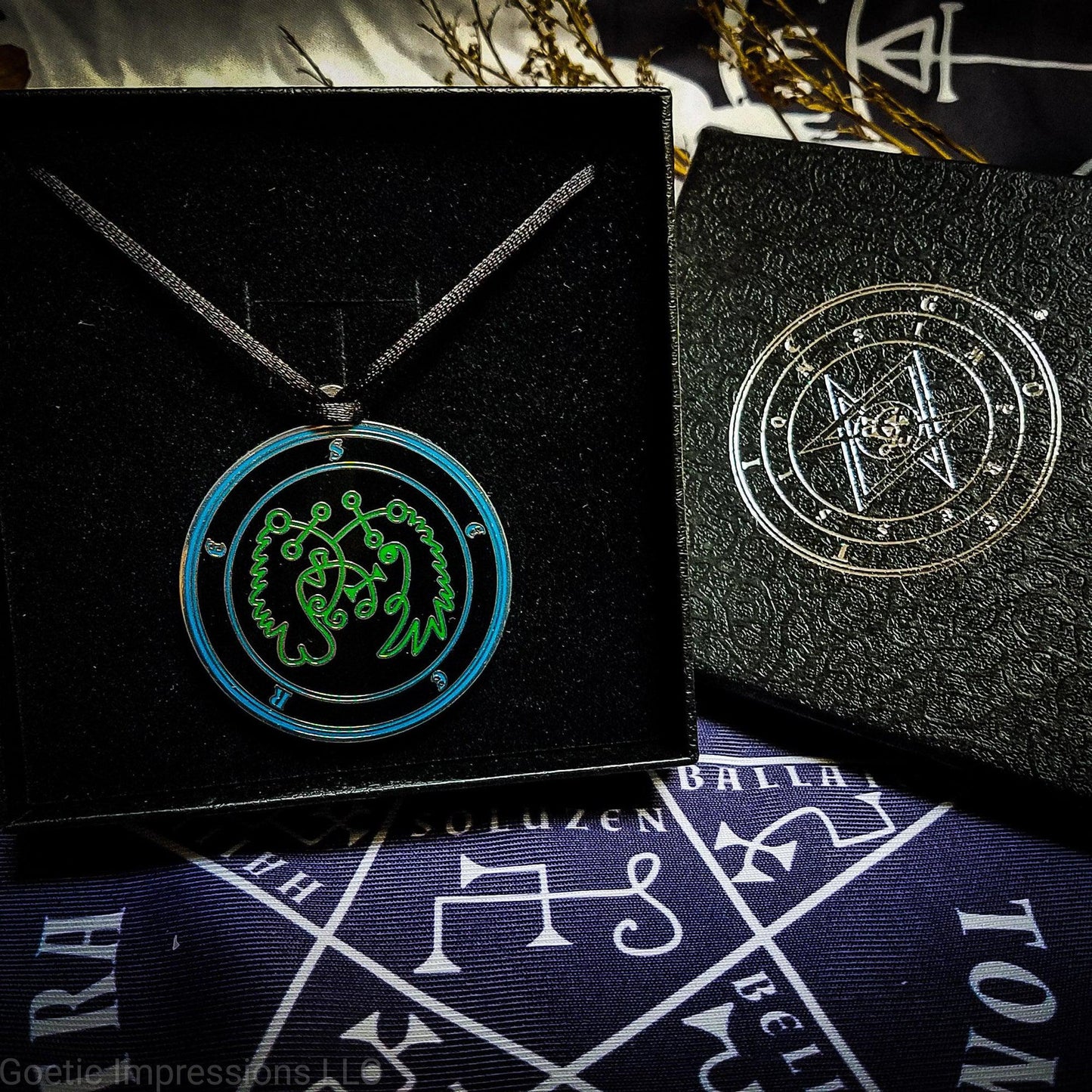 Necklace of Seere in a Goetic Impressions gift box stamped in silver foil. The sigil for Seere is green. Seere's name is surrounding the sigil with concentric circles in blue on a black background.  The seal is silver plated. The box is on a purple altar cloth with the Tetragrammaton in white.