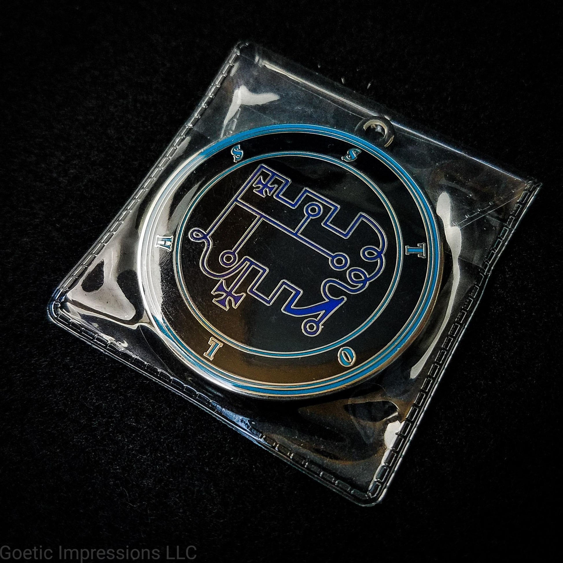 Amulet of Stolas in a PVC pouch. The sigil for Stolas is dark blue. Stolas' name is surrounding the sigil with concentric circles in teal on a black background. The seal is silver plated.