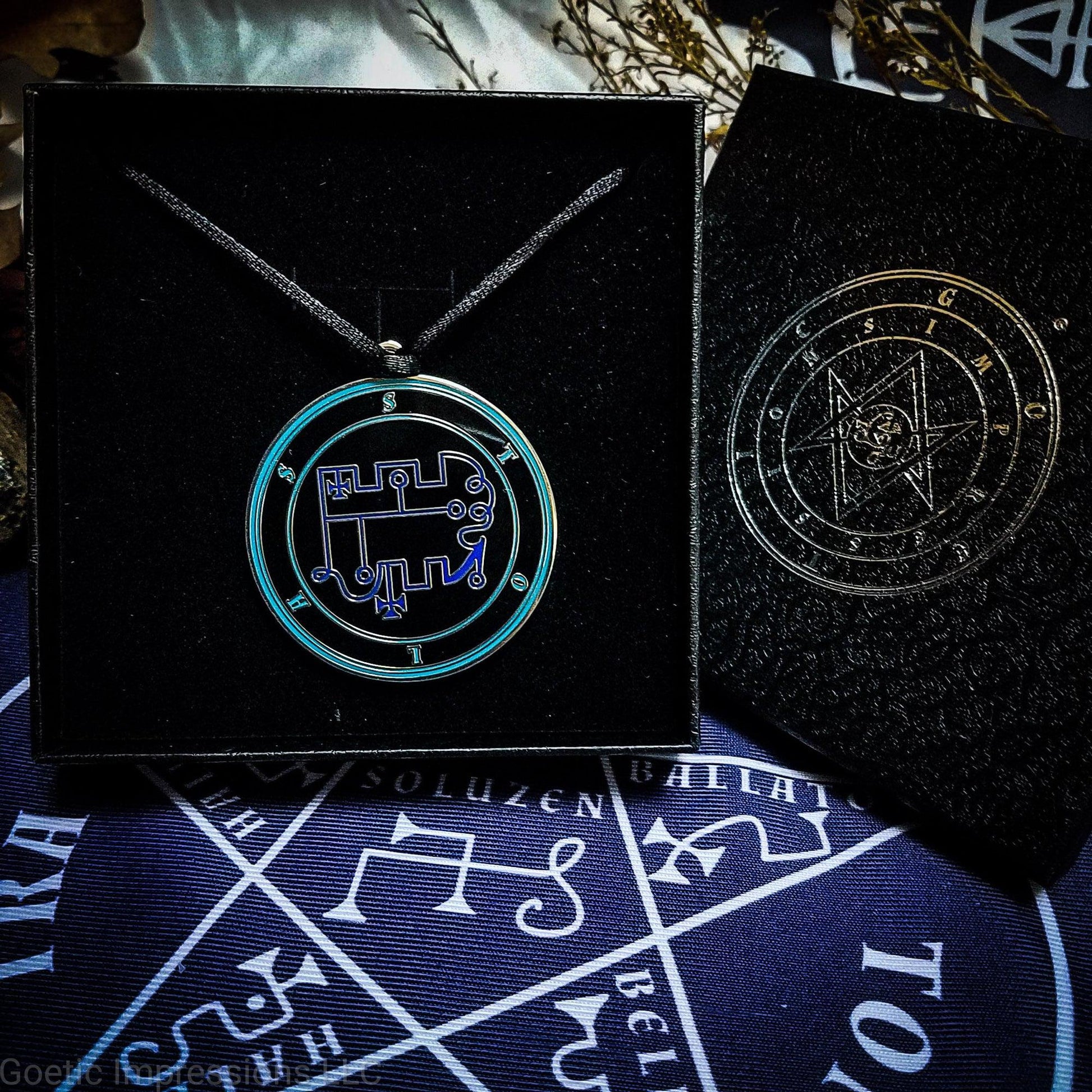 Necklace of Stolas in a Goetic Impressions gift box stamped in silver foil. The sigil for Stolas is dark blue. Stolas' name is surrounding the sigil with concentric circles in teal on a black background. The seal is silver plated. The box is on a purple altar cloth with the Tetragrammaton in white.
