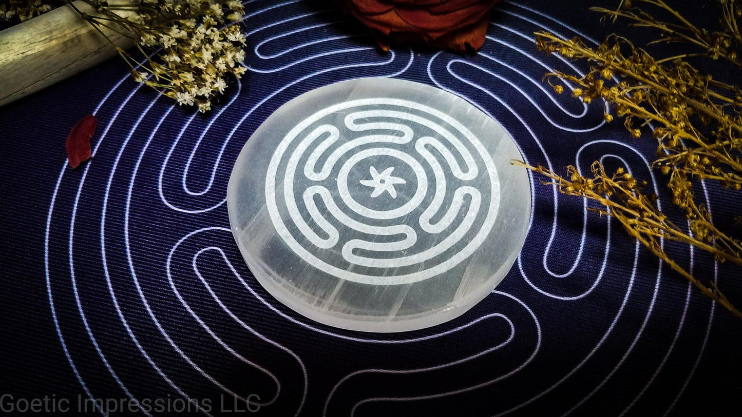 A selenite charging plate with the Wheel of Hecate or Stropholos engraved in it. The plate is placed on a purple altar cloth also with a white hecate sigil.